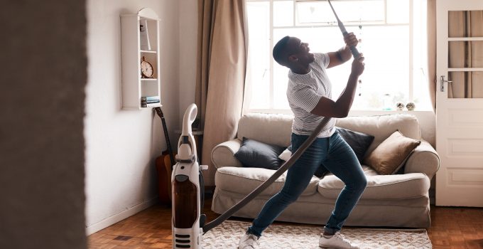 A young black man air guitars with the hoover whilst cleaning at home