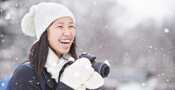 A woman in a hat and mittens holds a camera and laughs as it's snowing