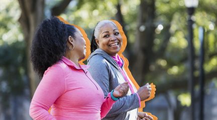 Two women exercising together in the city, jogging or power walking, laughing and conversing. Buildings and trees are out of focus in the background. The one in pink is in her 60s and her friend is in her 50s.