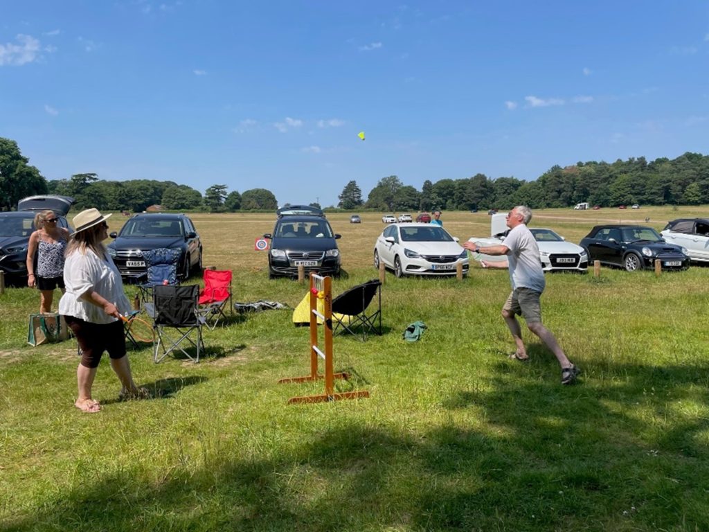 A group of people playing a bat and ball game on a sunny day in a park with cars parked in the background