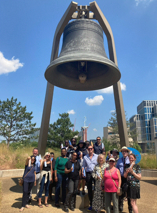 A diverse group of people stood beneath a large old bell on a sunny day in Stratford. They are stood on a grassy area with trees and buildings in the background. The bell is mounted on a metal frame with four legs and has a clapper hanging from the inside.